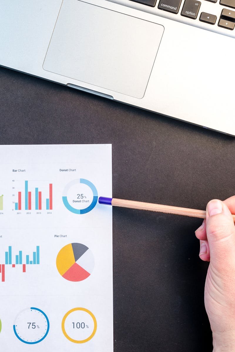 Hand holding pencil reviewing colorful data charts on desk with laptop.